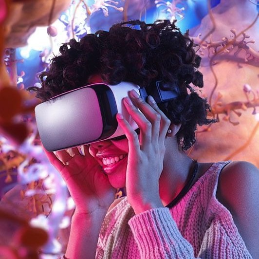 A smiling person with curly hair wears a virtual reality headset, surrounded by colorful, abstract lights and shapes, creating an immersive and futuristic atmosphere.